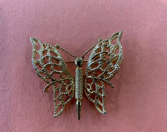 May include: A gold-toned butterfly brooch with intricate detailing. The wings are delicate and lacy, and the body is a long, slender pin.