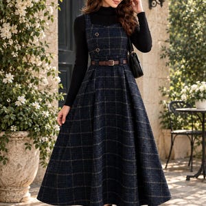 Puede incluir: Un vestido tipo pichi azul marino a cuadros con escote cuadrado, dos botones decorativos en el corpiño y un cinturón de cuero marrón en la cintura. El vestido se lleva sobre un suéter negro de cuello alto y manga larga con botines negros.