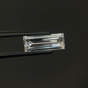 May include: A clear, rectangular baguette-cut gemstone with sharp, linear facets that reflect light. The stone is held horizontally by a pair of black metal tweezers, showcasing its elongated, transparent, and highly reflective crystalline structure.
