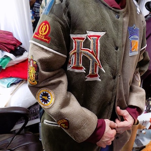 Peut inclure: Une veste bomber en daim vert avec un grand "H" brodé sur le devant. La veste comporte plusieurs patchs, dont un blason de Poudlard, un patch "Poufsouffle" et un patch "Association Naturelle de Quidditch".