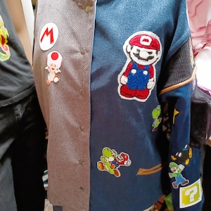 May include: A blue and gray varsity jacket with various embroidered patches featuring characters from the Super Mario Bros. video game franchise. The patches include Mario, Luigi, Yoshi, and a question mark block.