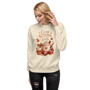 May include: A cream-colored crewneck sweatshirt featuring a graphic print of a mug filled with a latte, topped with whipped cream and cinnamon sticks, surrounded by autumn leaves, small pumpkins, and a plaid blanket with the text Cozy Autumn Latte.