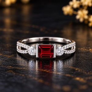 Könnte beinhalten: Ein silberfarbener Ring mit einem zentralen rechteckigen roten Edelstein, der von zwei runden weißen Steinen flankiert wird. Die Ringschiene ist geteilt und an den Seiten mit kleinen weißen Steinen besetzt, was für ein detailliertes Aussehen sorgt.