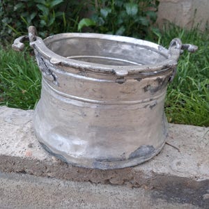 Peut inclure: Un seau en métal argenté avec une large ouverture et deux poignées latérales. La surface extérieure présente une texture martelée et patinée avec des signes d'usure visibles, incluant des zones décolorées et une base légèrement irrégulière.