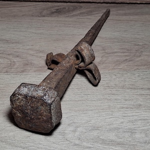 Op de afbeelding: Een handgesmede ijzeren spijker met een grote, vierkante kop en een taps toelopende, puntige schacht. Het metalen oppervlak vertoont een zware, roestbruine patina met een decoratief, gekruld metalen accent bevestigd nabij de kop.