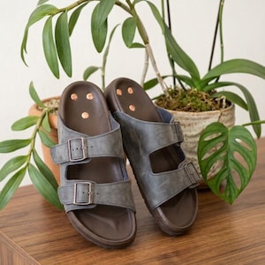 Peut inclure: Une paire de sandales grises à double bride avec des semelles marron foncé et trois petits rivets couleur cuivre au niveau du talon. Chaque bride est fixée par une boucle métallique, et le design inclut une semelle profilée.