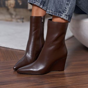 Peut inclure: Une paire de bottines marron foncé avec un bout pointu et un talon compensé. Les bottines sont fabriquées dans une matière lisse et présentent une fermeture éclair latérale. La tige monte au-dessus de la cheville, offrant une silhouette épurée.