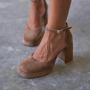 May include: A pair of brown suede platform shoes with a square toe, a thick block heel, and a thin ankle strap featuring a small silver-toned metal buckle. The shoes have a solid, uniform brown color and a soft, textured suede finish throughout.