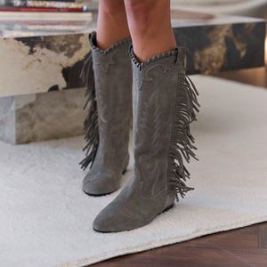 May include: Mid-calf grey suede boots featuring long fringe detailing along the outer sides, pointed toes, and decorative stitching patterns on the shafts. The boots have a flat sole and a scalloped edge finish at the top of the boot opening.