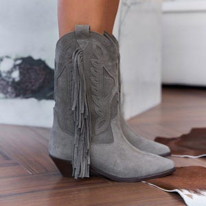 Könnte beinhalten: Ein Paar graue Wildleder-Cowboystiefel mit spitzer Zehenpartie, einem Blockabsatz und dekorativen Ziernähten am Schaft. Die Stiefel sind mit langen, passenden grauen Fransen an der Seite verziert und verfügen über eine Anziehlasche am oberen Rand.