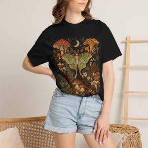 May include: A black t-shirt featuring a central graphic print of a large green moth surrounded by various mushrooms, ferns, and a crescent moon. The design includes red, orange, and brown fungi set against a dark background on the front of the shirt.