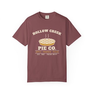 Puede incluir: Una camiseta de manga corta color granate con un gráfico de un pastel con vapor saliendo de él. El texto dice HOLLOW CREEK PIE CO. con las palabras APPLE, PUMPKIN, PECAN debajo, seguidas de EST. 1987 FRESH DAILY en tonos crema y amarillo.