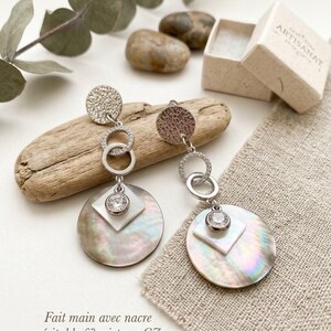 May include: A pair of silver-toned drop earrings featuring a textured circular top, two interlocking rings with small clear stones, and a large iridescent mother-of-pearl disc layered with a white square and a central clear crystal stone.