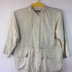 May include: A light beige hooded jacket featuring a full-length front zip, long sleeves, and a drawstring waist. The garment includes two zipped chest pockets and two lower flap pockets, all in a matching light beige fabric.