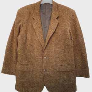 May include: A brown and tan tweed blazer featuring a textured houndstooth pattern, notched lapels, two front buttons, and two flap pockets. The garment is fully lined with a smooth brown interior fabric and is displayed on a plastic hanger.