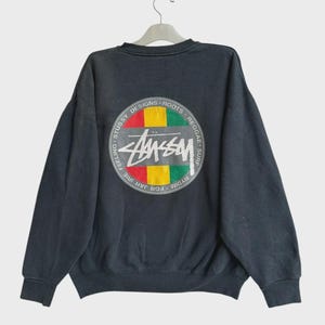 Op de afbeelding: Een donkergrijs sweatshirt met lange mouwen en een cirkelvormige afbeelding op de achterkant. De afbeelding bevat rode, gele en groene horizontale strepen, een wit Stussy-logo en de tekst Stussy Designs Roots Reggae Surf For Jah Irie Feeling.