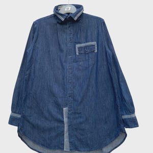 May include: A long-sleeve denim shirt in a medium blue wash featuring a button-down front, a pointed collar with light blue trim, a single chest pocket with a light blue flap, and a vertical light blue panel detail along the lower front hem.