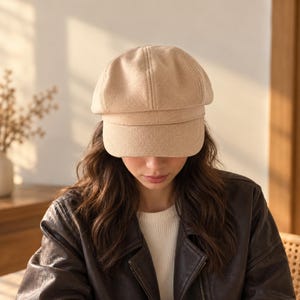 May include: A beige newsboy cap with a structured crown and a short, stiff brim. The cap features a textured fabric construction with visible vertical seams meeting at the top center, providing a classic rounded silhouette and a clean, solid finish.