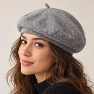 May include: A soft, light gray knit beret featuring a classic rounded silhouette with a small, centered top knot detail. The hat is constructed from a fine-gauge, textured wool-blend fabric that provides a structured yet flexible shape.