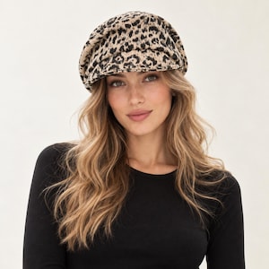 May include: A newsboy cap featuring a tan and black leopard print pattern. The hat has a rounded, voluminous crown and a short, stiff brim at the front. The fabric displays a textured appearance with a repeating animal spot design.