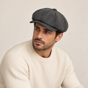 May include: A dark gray wool newsboy cap featuring an eight-panel construction with a central button at the crown and a short, stiffened brim. The cap has a textured fabric finish and a rounded, structured shape that sits firmly on the head.