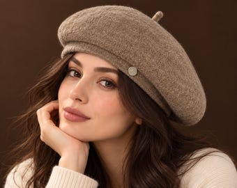 May include: A light brown beret with a textured, felted appearance and a small, circular metallic accent on the side. The hat features a classic rounded crown with a small center tip, providing a soft, uniform color and a structured yet pliable shape.