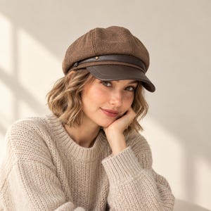 May include: A brown newsboy cap featuring a textured fabric crown and a smooth dark brown faux leather brim. A thin matching faux leather strap runs across the base of the crown, secured by a small metallic stud on the side of the hat.