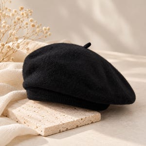 May include: A solid black beret featuring a soft, fuzzy texture and a small, centered top knot. The hat has a structured, ribbed knit band at the base, providing a clean, circular silhouette for this classic headwear accessory.