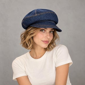 May include: A dark blue denim newsboy cap featuring a structured crown, a short stiff brim, and visible copper-colored stitching along the seams. The hat is crafted from a durable fabric with a classic multi-panel construction and a rounded shape.