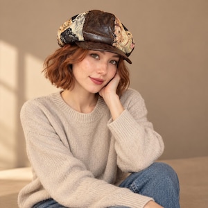 May include: A newsboy cap featuring a dark brown leather center panel and side panels with a multi-colored floral tapestry pattern. The hat has a structured brim and a rounded crown, constructed from a combination of smooth leather and woven fabric.