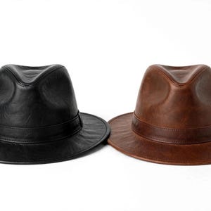Puede incluir: Dos sombreros fedora, uno en negro sólido y otro en marrón medio, ambos con corona pellizcada, ala ancha y una banda a juego de aspecto cuero alrededor de la base. Los sombreros tienen un acabado liso y ligeramente texturizado.