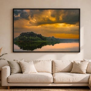 May include: A rectangular landscape wall art print in a thin black frame featuring a sunset over a calm lake with dark clouds and a treeline. The artwork is displayed above a beige sofa with matching throw pillows and a textured throw blanket.