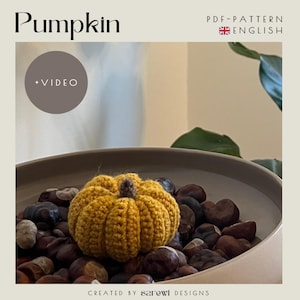 Könnte beinhalten: Ein senfgelber gehäkelter Kürbis mit einem kleinen braunen Stiel, der in einer Schale mit dunkelbraunen Kastanien liegt. Der Text lautet Pumpkin, +VIDEO, PDF-PATTERN ENGLISH und CREATED BY sarewi DESIGNS.