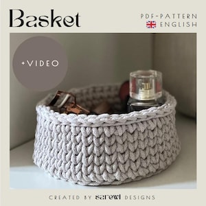 May include: A light gray crocheted basket with a thick, textured stitch pattern. The text "Basket", "+ VIDEO", "PDF-PATTERN ENGLISH", and "CREATED BY sarewi DESIGNS" is visible. Inside the basket sits a perfume bottle and a metal eyelash curler.