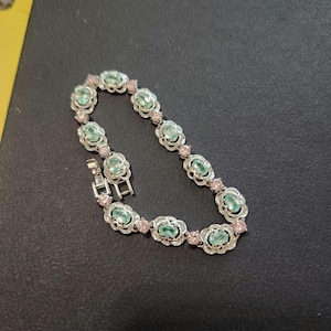 Puede incluir: Una pulsera de tono plateado que presenta una serie de piedras ovaladas de color verde claro engastadas en marcos metálicos decorativos, alternadas con pequeñas piedras redondas de color rosa. Incluye un cierre de caja metálico seguro.