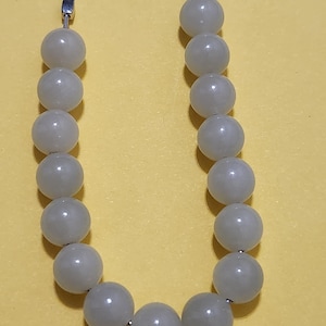 Puede incluir: Un collar que presenta una sola hilera de cuentas redondas y lisas de color blanco roto. Las cuentas están ensartadas en una fina cadena de metal plateado, con pequeños espaciadores de metal plateado en los extremos.
