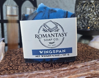 Può includere: Una saponetta rettangolare blu con un'etichetta di carta bianca riportante il testo Romantasy Soap Co. Wingspan e un peso netto di 4,5 oz | 127 g. Il sapone ha un aspetto marmorizzato blu scuro e nero con una superficie superiore ruvida.