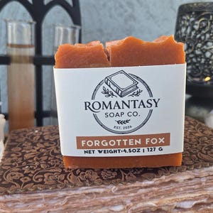 Puede incluir: Una pastilla de jabón rectangular de color naranja envuelta en una etiqueta de papel blanco. La etiqueta incluye el texto ROMANTASY SOAP CO., FORGOTTEN FOX y NET WEIGHT 4.5OZ | 127 G, junto a un logotipo circular con un libro y ramas.