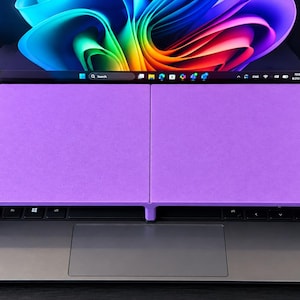 May include: A purple laptop keyboard cover stand resting on a dark gray laptop. The stand features a flat, rectangular surface divided into two sections, positioned directly over the keyboard area, with a vibrant abstract wallpaper on the screen.