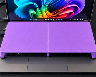 Könnte beinhalten: Ein violetter Laptop-Tastaturständer auf einem dunkelgrauen Laptop. Der Ständer verfügt über eine flache, rechteckige Oberfläche, die in zwei Abschnitte unterteilt ist und direkt über dem Tastaturbereich liegt, mit einem abstrakten Hintergrundbild.
