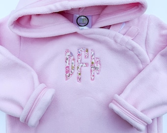 May include: A light pink fleece hoodie featuring a wrap-style neckline and long sleeves. The center of the chest displays a three-letter monogram in a floral fabric pattern. A white tag inside the neck area shows the text BB and 2T.
