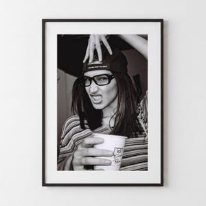 Peut inclure: Une impression encadrée en noir et blanc montrant une personne portant une casquette avec le texte YOU ARE WHAT YOU DRIVE, des lunettes à monture foncée et un haut rayé à manches longues, tenant un gobelet blanc avec un motif et le mot Pain.
