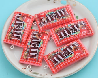 May include: Five pink and red M&M's candy bar charms with silver jump rings. The charms are shaped like a candy bar wrapper with the M&M's logo and the words "Milk Chocolate" printed on them.