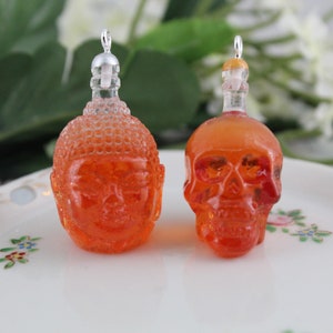 May include: Two small glass bottle pendants, one shaped like a Buddha head and the other shaped like a skull. Both pendants are filled with a clear orange liquid.