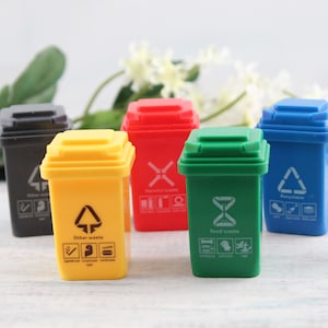 May include: Five miniature trash cans in different colors, including black, yellow, red, green, and blue. Each can has a different recycling symbol and label, such as "Other waste", "Food waste", and "Recyclable".