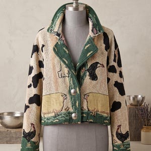 May include: A cropped jacket featuring a woven tapestry design with farm animals including sheep and roosters on a cream background. The jacket has a green collar and trim, long sleeves with black cow-like spots, and two silver-toned metal buttons.