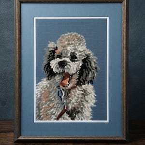 Puede incluir: Un retrato en punto de cruz enmarcado de un perro esponjoso blanco y negro con la boca abierta, sobre un fondo azul. La obra está en un marco de madera oscura con paspartú azul, mostrando puntadas detalladas del pelaje canino.