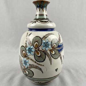 May include: A ceramic vase with a cream-colored base featuring hand-painted floral motifs in shades of light blue, sage green, and dark brown. The design includes intricate fern-like leaves and small blossoms, with a distinct blue band encircling the neck.