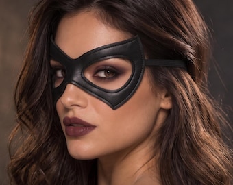 Könnte beinhalten: Eine schwarze Augenmaske in Lederoptik mit spitzer, katzenohrähnlicher Silhouette und einem dünnen elastischen Band. Die Maske hat glatte, vernähte Kanten und bedeckt den Augenbereich, während Nase und Mund für ein passgenaues Aussehen frei bleiben.