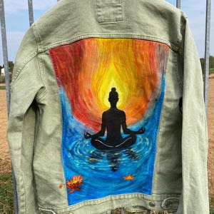 Könnte beinhalten: Eine olivgrüne Jeansjacke mit einem handgemalten Design auf der Rückseite. Das Kunstwerk zeigt die schwarze Silhouette einer Person in sitzender Meditationshaltung, umgeben von einem leuchtend orange-gelben Flammenmotiv und blauem Wasser.